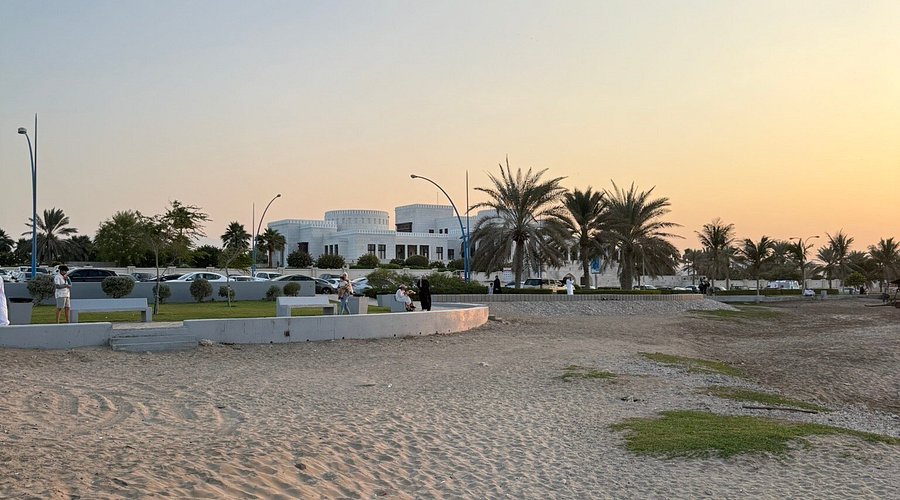Sunset over Al Azaiba Beach Park in Muscat, Oman, featuring sandy shores, palm trees, stone walkways, and white modern residential villas in the background.