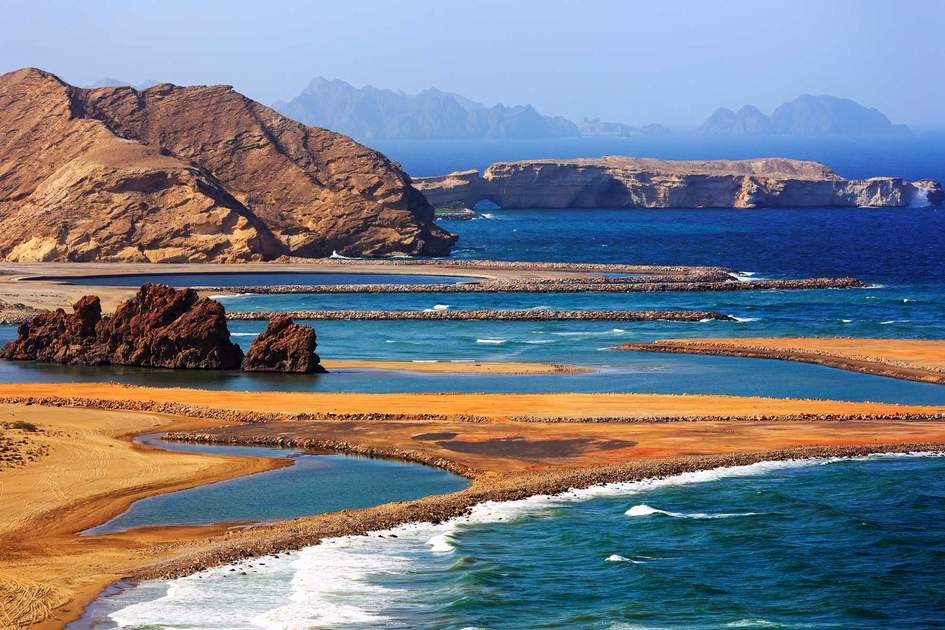 Aerial view of the dramatic rocky coastline at Bandar Al Jissah and Yiti Beach in Muscat, Oman, featuring clear blue lagoons, golden sand banks, and a natural stone arch in the distant sea cliffs.