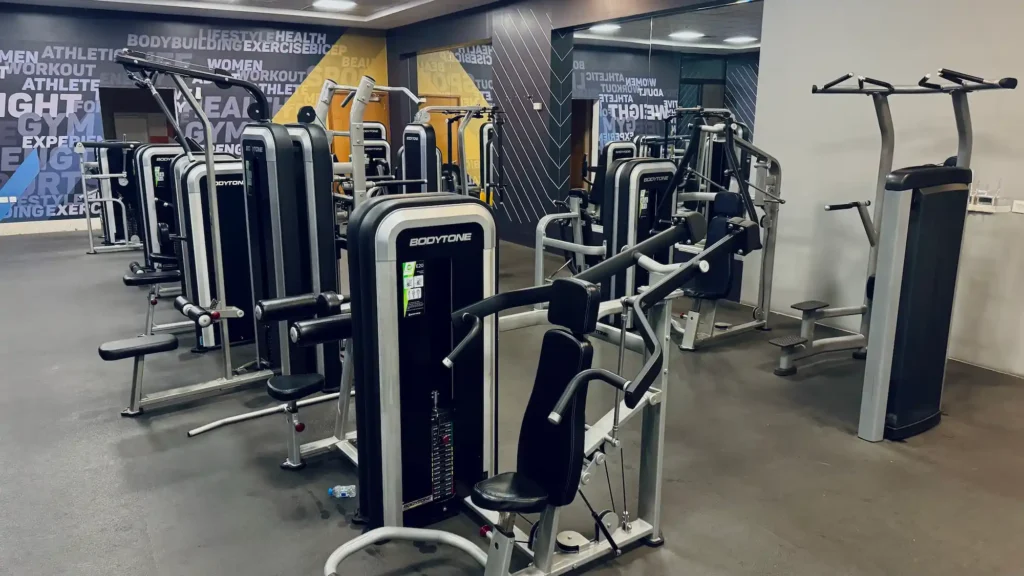 Modern gym in Muscat with cardio machines and strength equipment Strength training equipment and free weights at a gym in Muscat Cardio machines for weight loss and fitness training in Muscat gym Personal trainer helping a gym member with exercise technique in Muscat Clean and modern fitness gym in Oman for beginners and athletes Gym member training with free weights in Muscat