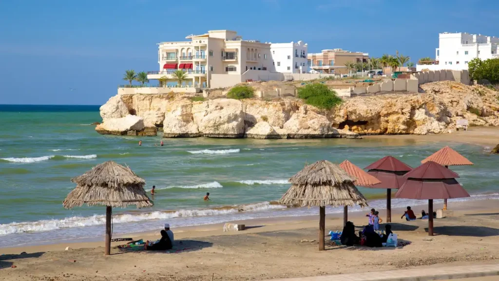 People relaxing on a sandy beach under thatched umbrellas with white luxury villas sitting on a rocky cliff overlooking the turquoise sea.