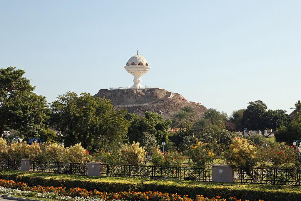 The giant white Riyam Park Incense Burner monument on a rocky hill overlooking lush green gardens and flowers in Muttrah, Muscat, Oman